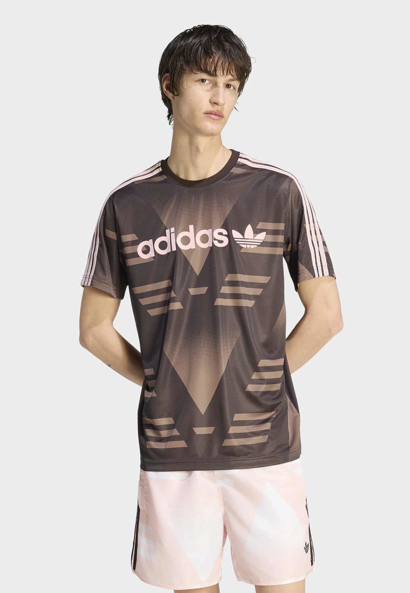 Giovane uomo che indossa una maglietta marrone a motivi Adidas e pantaloncini rosa chiaro con le mani dietro la schiena, davanti a uno sfondo chiaro e uniforme.