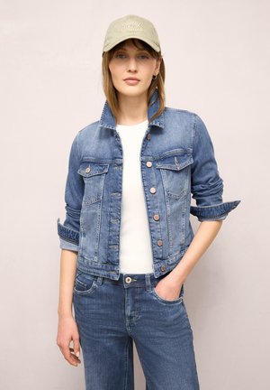 Kvinde iført beige baseballkasket, blå denimjakke med opkrogede ærmer, hvid skjorte og blå jeans, poserer med den ene hånd i lommen.