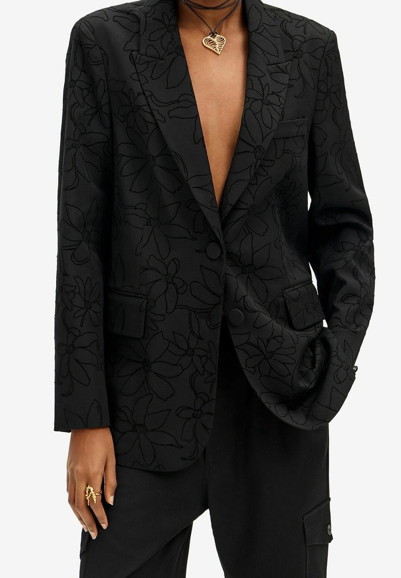 Personne portant un blazer noir à motif floral avec un profond décolleté, un pantalon noir, un collier en or en forme de cœur et une bague dorée remarquable.