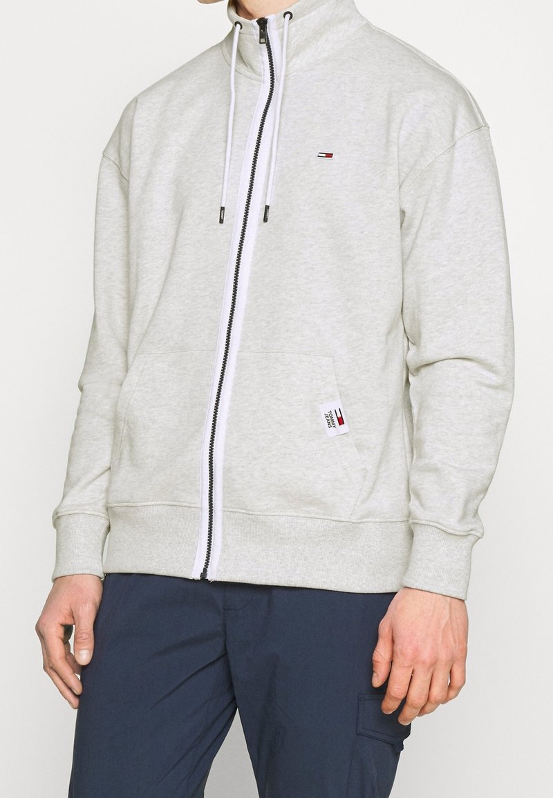 Homme portant un sweat à capuche gris clair zippé avec des cordons blancs et un petit logo sur la poitrine et la poche, associé à un pantalon bleu marine.