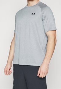 Mężczyzna ubrany w jasnoszary, krótki rękaw, sportowy T-shirt z czarnym logiem Under Armour na klatce piersiowej oraz czarne spodenki sportowe.