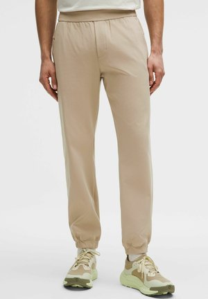 Man die beige joggers met elastische tailleband en beige- en gebroken-witte sneakers draagt, staand tegen een effen lichte achtergrond.
