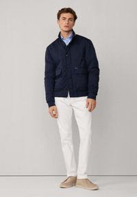 Chaqueta acolchada azul marino con cuello alto, botones en el frente, dos bolsillos con solapa, combinada con pantalones blancos y zapatos beiges. Textura de tela suave.