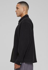 Black button-up jacket with a relaxed fit, textured fabric, wide sleeves, and a collar. Features a silver button detail at the cuff.