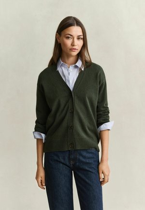 Vrouw die een donkergroene, met knopen gesloten cardigan draagt over een lichtblauw overhemd met opgerolde mouwen en donkerblauwe spijkerbroek, staand tegen een effen achtergrond.