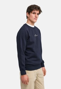 Marineblå genser i bomullsblanding, med rund hals og ribbestrikkede mansjetter. Liten hvit logo på brystet. Glatt tekstur, avslappet passform.