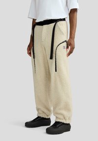 Kremowe spodnie dresowe z fakturowanym wykończeniem, z czarnym paskiem i zamkami błyskawicznymi po bokach; zestawione z czarnymi sneakersami.