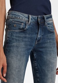 Jeans de mezclilla en un lavado azul oscuro descolorido, con un diseño estándar de cinco bolsillos, herrajes de metal y una textura ligeramente desgastada.