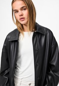 Schwarze Kunstleder-Übergrößejacke mit spitzem Kragen, Knopfleiste und glatter Textur, kombiniert mit einem weißen Oberteil darunter.