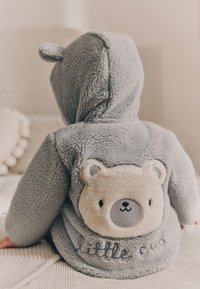 Pelēka, samtainā kapuce ar lāča ausīm uz kapuces, lāča uzlīme uz muguras un teksts "mazais lācis" izšūtu detaļu formā.