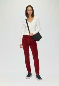 Langärmliges, weißes, geripptes Oberteil mit Knöpfen, kombiniert mit roten Cordhosen und schwarzen sportlichen Schuhen. Eine schwarze Umhängetasche rundet das Outfit ab.