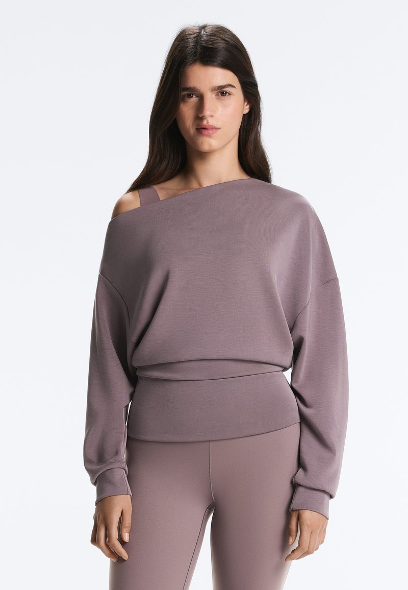 Vrouw draagt een mauve off-shoulder top met lange mouwen en zichtbare band, gecombineerd met strakke bijpassende broek, staand tegen een neutrale lichte achtergrond.