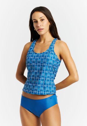 Vrouw die een blauw patroon tanktop en effen blauwe zwemshorts draagt, met haar handen achter haar rug staand tegen een witte achtergrond.
