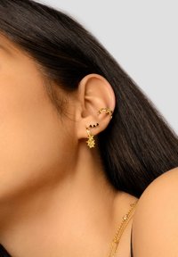 Orecchini d'oro con accenti a forma di stella e perline nere, design a catena texturizzata, indossati su un orecchio con capelli scuri e lisci.
