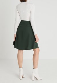 Jupe A-line verte faite d'un tissu lisse, avec une taille haute et un design fluide. Portée avec des bottines blanches, sur un fond uni.