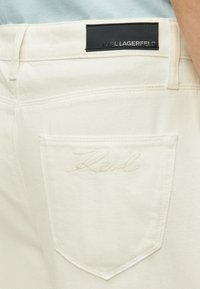 Κρεμ τζιν παντελόνια με μαύρη δερμάτινη ετικέτα με την επιγραφή "KARL LAGERFELD" και κεντημένη υπογραφή στην πίσω τσέπη.