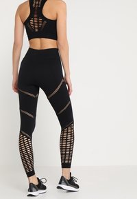 Černý sportovní crop top s otevřeným síťovaným zádovým dílem; odpovídající černé legíny s vysokým pasem, které mají výřezové vzory podél nohavic; doplněné černými teniskami.