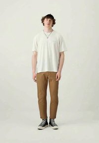 T-shirt beige, chinos marroni e sneakers neri. Maniche corte, vestibilità rilassata. Capelli ricci, in piedi su uno sfondo neutro. Abbigliamento casual.