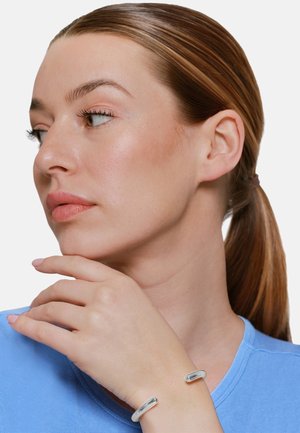 Donna con capelli castano chiaro raccolti in una coda di cavallo, indossa un top blu e un braccialetto aperto d'argento, guarda verso sinistra con la mano vicino al mento.