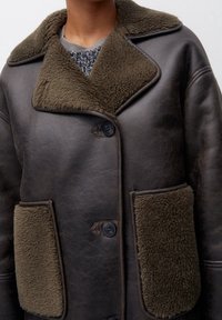 Abrigo de cuero marrón con un cuello de borreguillo texturizado en color verde oliva y acentos en los bolsillos. Presenta dos bolsillos en la parte delantera y cierre con botones.