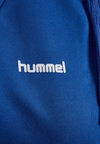 Blått tyg med ribbad textur som har en vit "hummel"-logotyp tryckt framträdande i mitten. Materialet verkar mjukt och slätt.