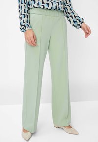 Hellgrüne weite Hose mit glatter Textur und elastischem Bund, kombiniert mit einer gemusterten Bluse und beigen spitzen Ballerinas.