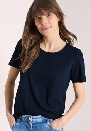 Jeune femme aux longs cheveux bruns portant un t-shirt bleu marine à manches courtes et un jean en denim bleu clair, les mains dans les poches.