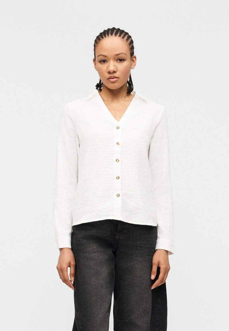 Femme aux cheveux tressés portant une chemise blanche à manches longues boutonnée et un jean noir délavé, debout devant un fond blanc uni.