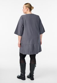 Šedé šaty s širokými rukávy a plisovaným lemem, vyrobené z hladkého materiálu. Spárované s vysokými černými botami a neprůhlednými punčochami.