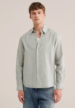 Ung mand med krøllet hår iført en lysegrå skjorte med knapper over en hvid undertrøje og blå jeans, stående mod en ensfarvet baggrund.