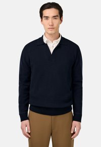 Námornícky pletený pulóver s výstrihom do V a štruktúrovaným golierom, s dlhými rukávmi a rebrovaným lemom. Nosený cez bielu košeľu s golierom.