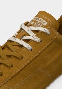 Κοντινό πλάνο καφέ σουέντ αθλητικού παπουτσιού Converse Star με λευκές και καφέ ραφές, με αντίθετα λευκά κορδόνια πάνω από τη γλώσσα.