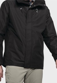 Muž oblečený v černé kapucové bundě se stahovacími šňůrkami a béžových kalhotách se zipovou kapsou, stojící na jednobarevném pozadí.