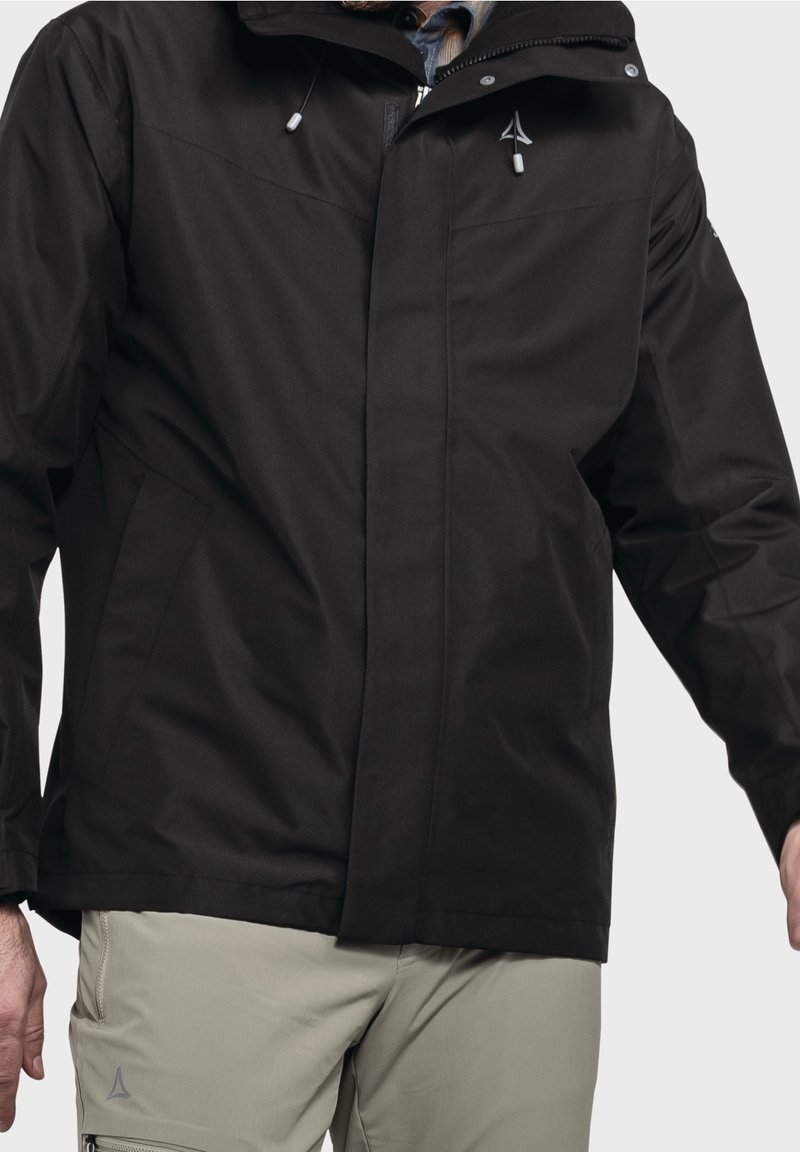 Muž oblečený v černé kapucové bundě se stahovacími šňůrkami a béžových kalhotách se zipovou kapsou, stojící na jednobarevném pozadí.