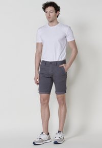 Férfi áll egy fehér háttér előtt, fehér pólóban, szürke felhajtott nadrágban és fehér sportcipőben, az egyik kezét a zsebébe dugva.