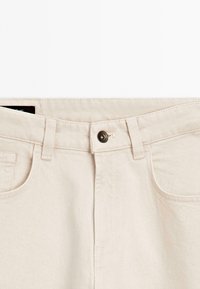 Nahaufnahme von beigen Denim-Jeans mit Knopfverschluss, Gürtelschlaufen und Fronttaschen.