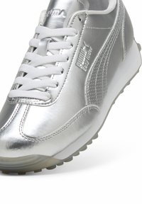 Scarpa atletica argento con superficie metallica lucida, lacci bianchi, dettagli texturizzati sui lati e suola in gomma arrotondata.