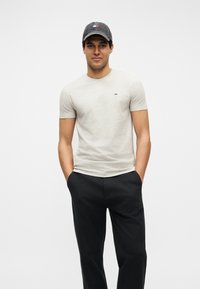 Άνδρας που φοράει ανοιχτό μπεζ εφαρμοστό κοντομάνικο μπλουζάκι, μαύρο παντελόνι και σκούρο γκρι καπέλο του μπέιζμπολ με τα χέρια στις τσέπες.