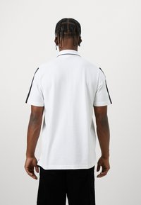 Camisa polo branca com detalhes laterais pretos, mangas curtas e colarinho clássico. O tecido parece suave. Usada sobre calças pretas.