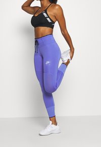 Fioletowe legginsy z teksturowanym wzorem i czarnymi akcentami, zestawione z czarnym stanikiem sportowym z logo. Całość dopełniają białe sneakersy.