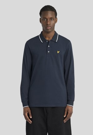 Jeune homme portant un polo bleu marine à manches longues avec des bordures blanches au col et aux poignets, debout devant un fond gris uni.