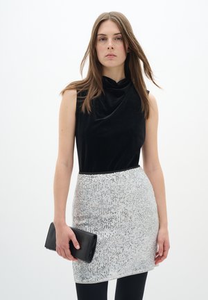 Schwarzes, ärmelloses Top aus Samt mit Wasserfallkragen, kombiniert mit einem silbernen Pailletten-Minikleid und einer schwarzen Clutch-Handtasche.