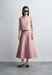 Moteris dėvi rožinį be rankovių viršutinį drabužį ir atitinkamą A formos sijoną, juodus katės akių formos saulės akinius ir juodus batelius su smailiu priekiu, stovinčią prie pilkos sienos.