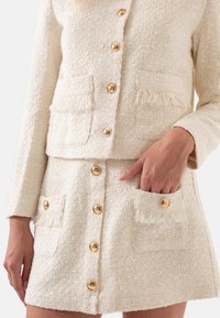 Crème tweed jasje en rok set met gouden knopen, franjes op de zakken en een gestructureerde stof. Met lange mouwen en een getailleerd ontwerp.