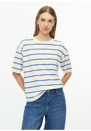 Femme aux cheveux châtain clair et raides porte un t-shirt blanc à rayures horizontales bleues et un jean bleu, debout les mains dans le dos.