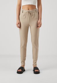 Beige gestreepte broek met een elastische taille en een trekkoord. Voorzien van een taps toelopend ontwerp en een gladde textuur. Draagt met zwarte sandalen.