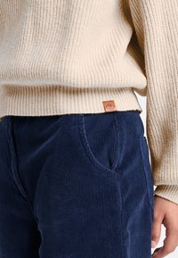 Beige ribbat tröja med en läderlogotypetikett, ihop med marinblå kordbyxor med ett strukturerat mönster och sidofickor.