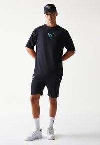 Μαύρο βαμβακερό μπλουζάκι με γραφικό ταύρο σε τιρκουάζ, συνδυασμένο με μαύρο σορτς που φέρει το ίδιο λογότυπο. Ολοκληρώνεται με μαύρο καπέλο και λευκά αθλητικά παπούτσια.