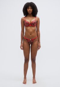 Σετ εσώρουχα με κόκκινο και μαύρο λεοπάρ σχέδιο που περιλαμβάνει σουτιέν με ενίσχυση και ρυθμιζόμενες τιράντες, καθώς και εσώρουχο τύπου σέξι σλιπ με χαμηλό καβάλο και διακοσμητικά μεταλλικά στοιχεία.