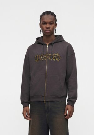 Giovane uomo con capelli intrecciati che indossa una felpa con cappuccio scura con zip con scritte gialle a motivo e jeans scuri scoloriti, in piedi davanti a uno sfondo semplice.